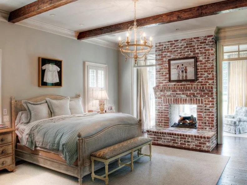 Tall red brick fireplace chimney in bedroom with wood beams, pale blue bed and bench