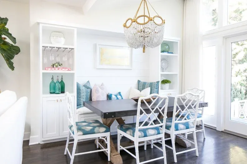 White fitted bench framed by shelving towers and lower cupboards, with blue pillows, a dark dining table, white lattice-back chairs, and a beaded chandelier.