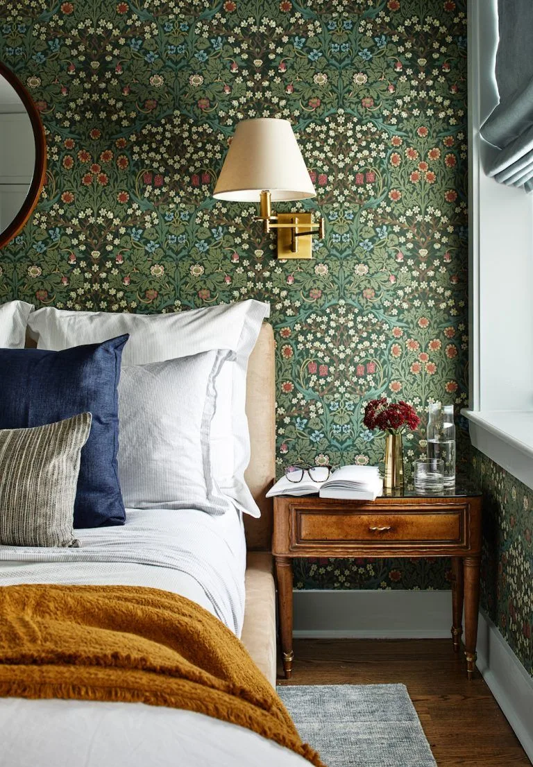 Dense green floral wallpaper behind pale bedding, a wooden bedside table and a cream shaded wall light.