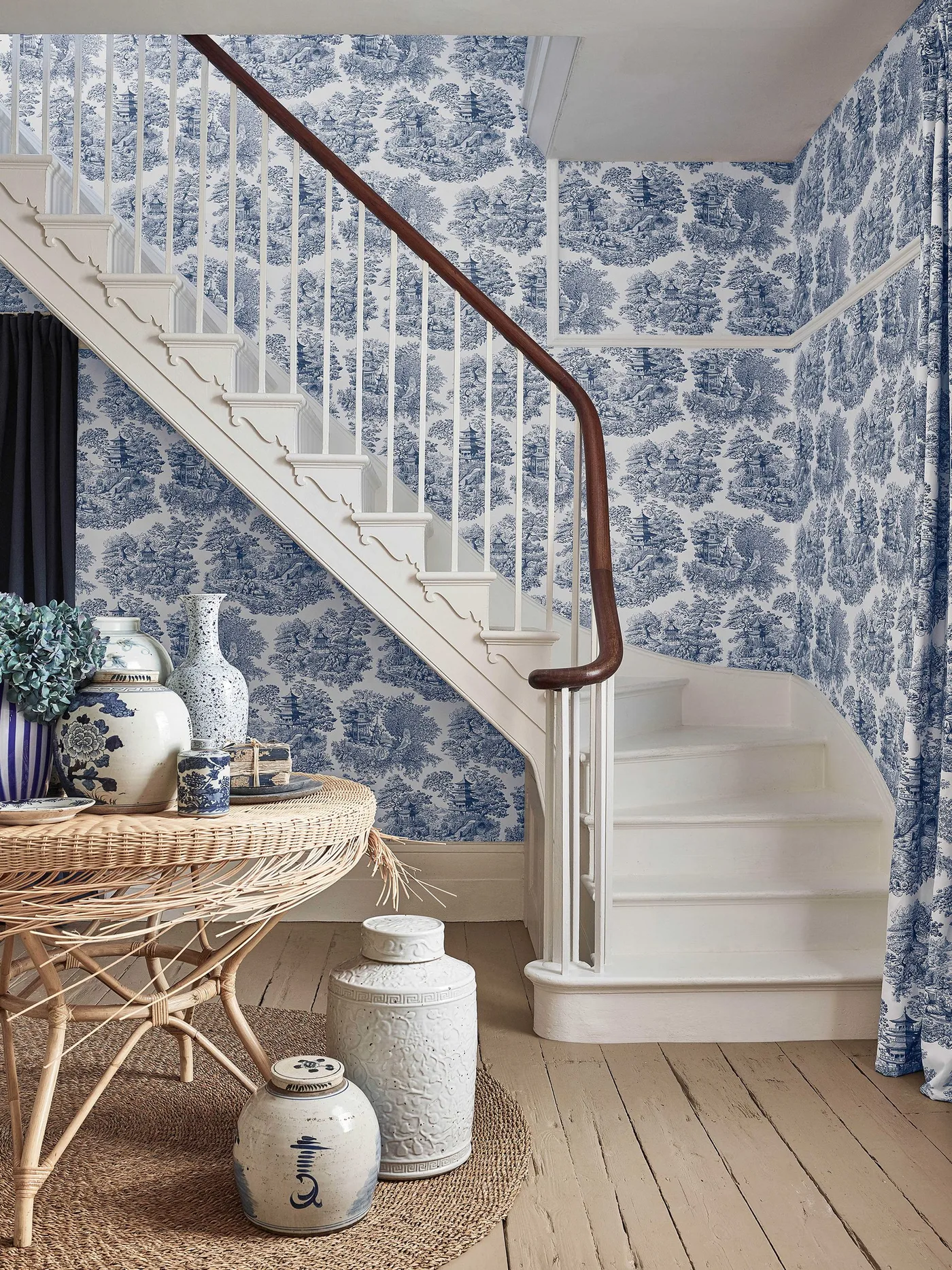 Blue-and-white patterned stair hall with a white staircase, wooden handrail and round woven table.
