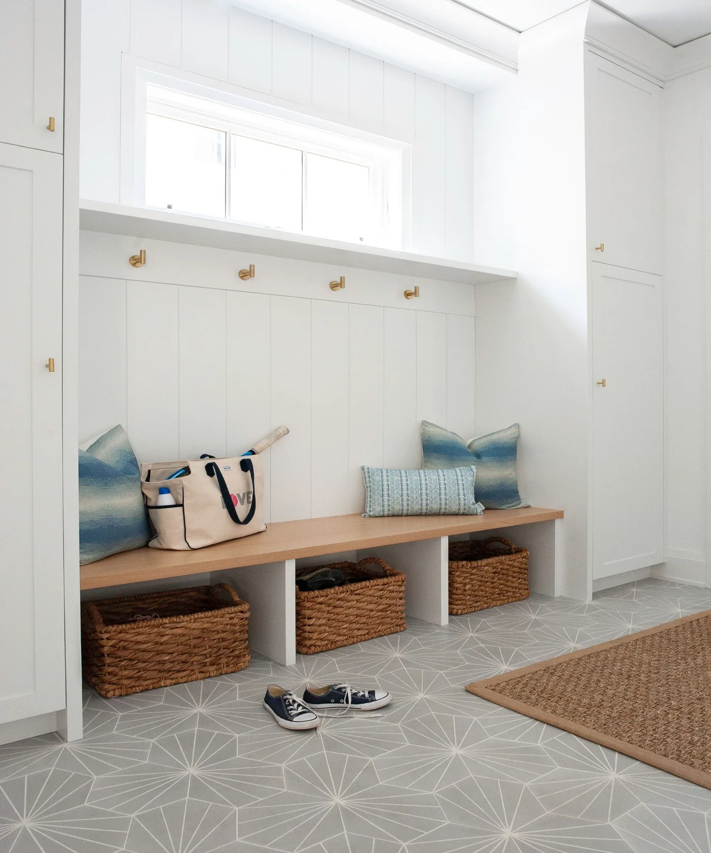 White mudroom with high windows, wood bench, three lower woven baskets, wall hooks, blue cushions and pale geometric floor