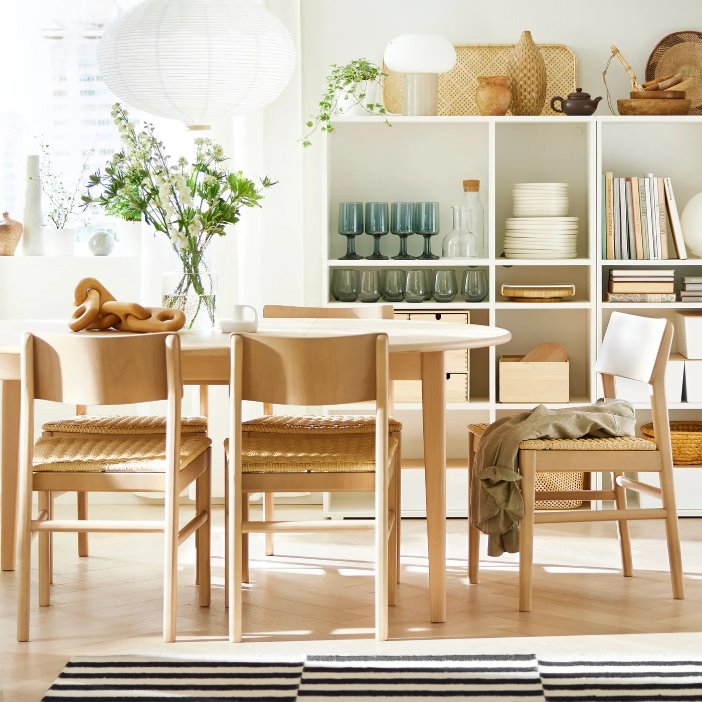 Pale wooden chairs with woven seats surround a round table beneath a white pendant beside white cubby shelves and a striped rug