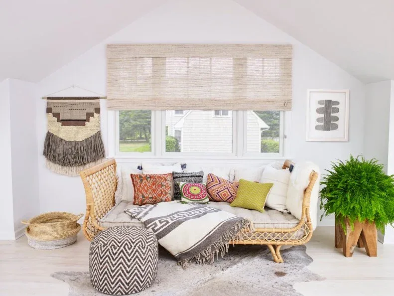 Woven settee beneath a window with colorful cushions, a fringed wall hanging, patterned pouf and fern.