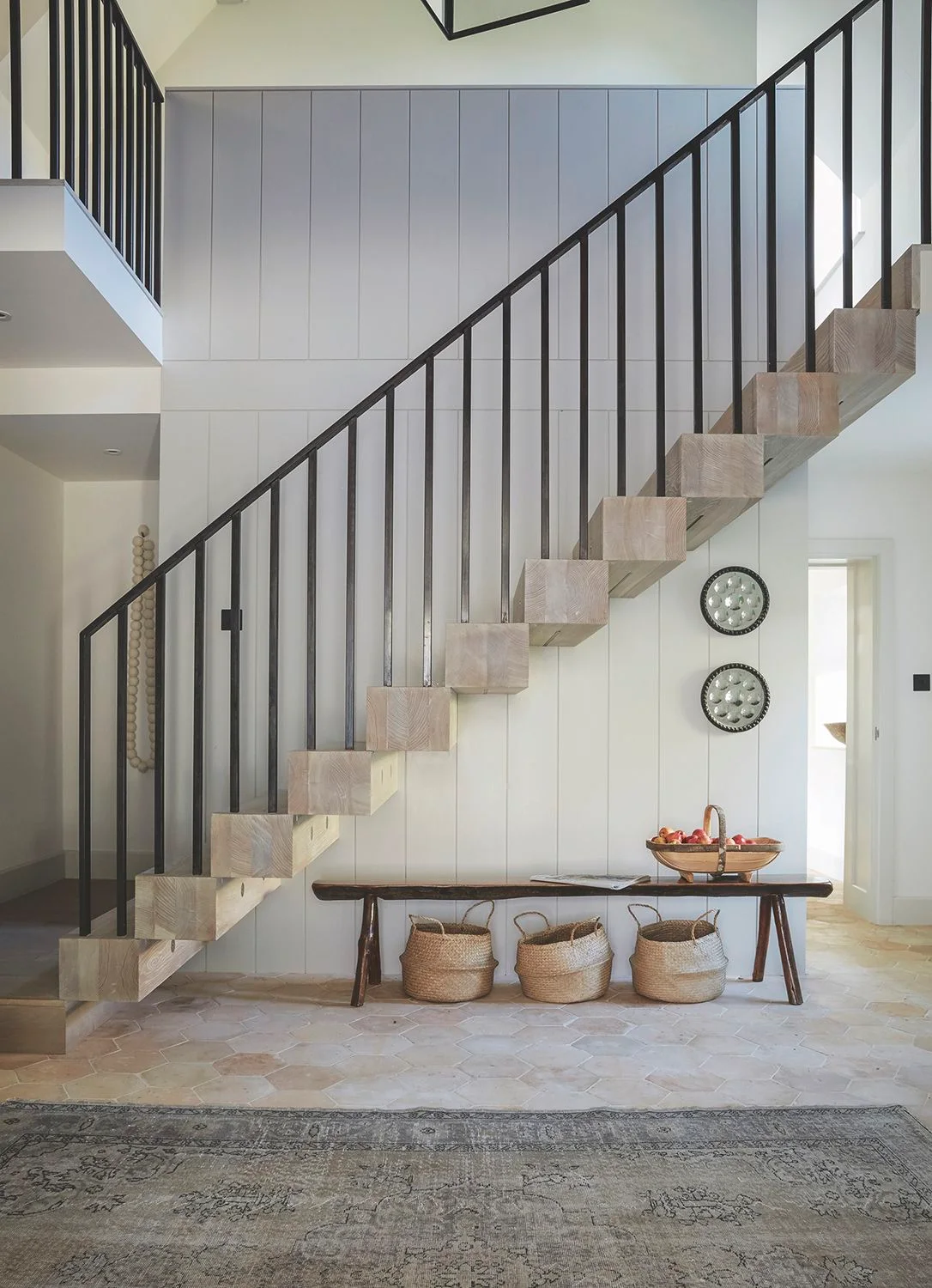 A pale entrance has black stair balusters above warmer steps and a wood bench with three baskets beneath beside a gray patterned rug