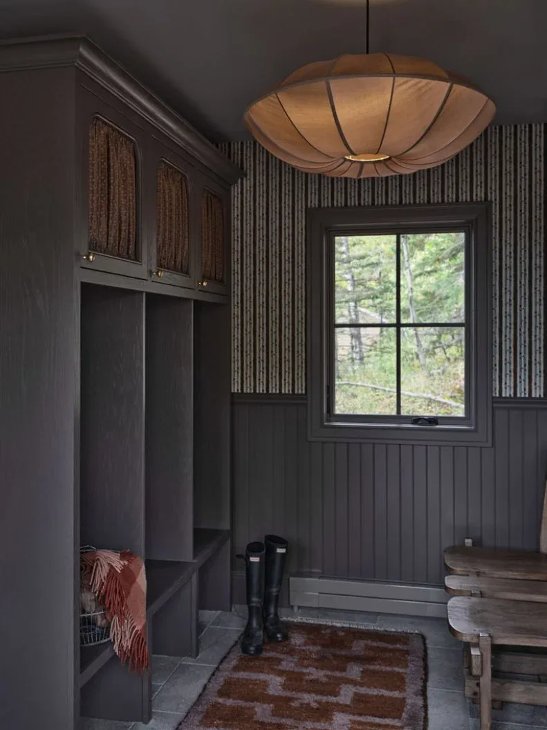 Dark gray mudroom with textured cabinet panels, wooden bench, patterned rug, fringed throw and broad pale pendant.