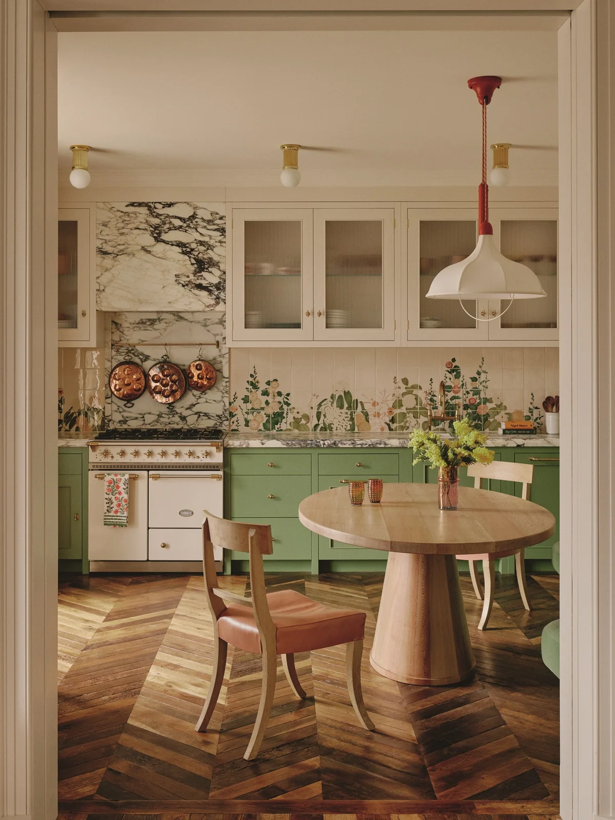 A green botanical scene on pale backsplash tiles sits beneath pale upper cupboards above green lower cabinets beside a round dining table