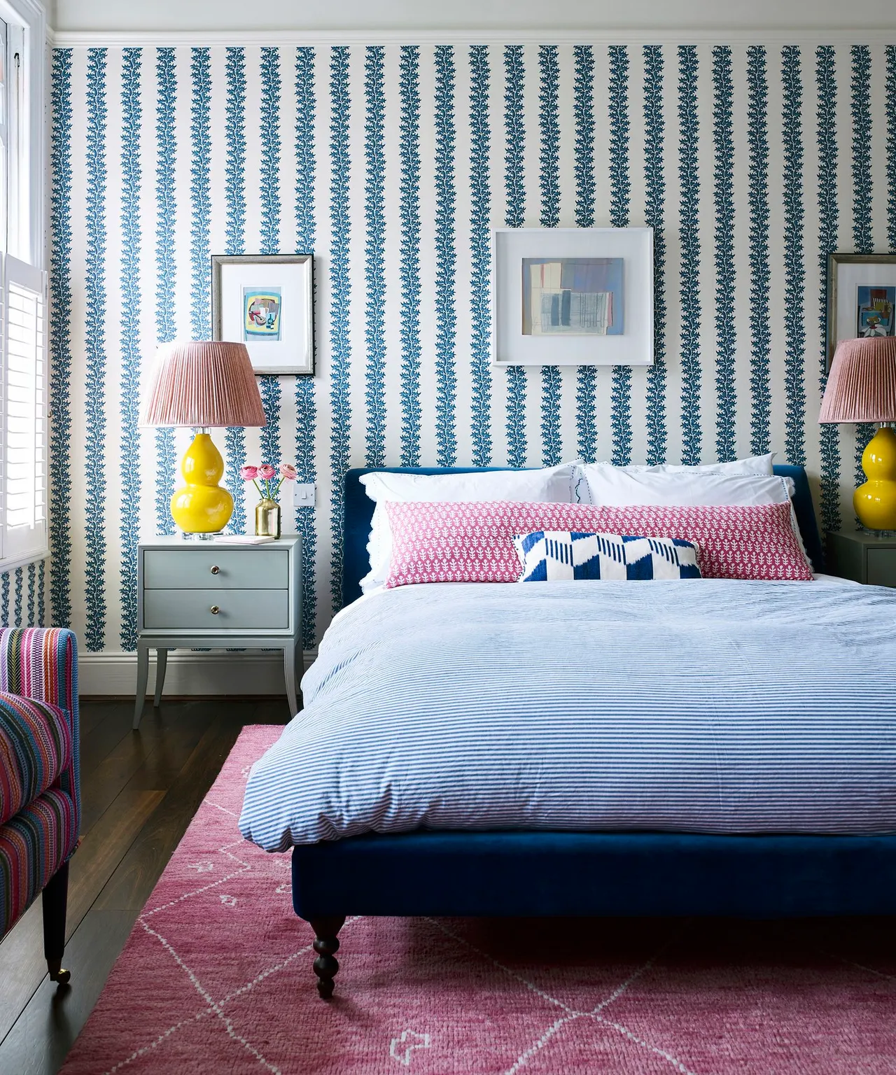 Bedroom with blue patterned vertical wallpaper, dark blue bed, pale bedding, pink rug, yellow lamp bases and gray-green bedside cabinets.