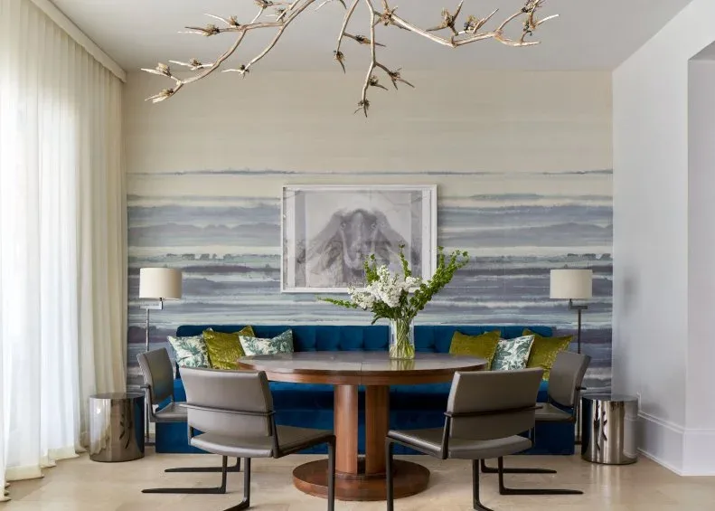 Dining nook with a blue-gray horizontal mural behind a blue banquette, framed picture, round wood table, gray chairs, metal floor lamps, and branch-like chandelier.