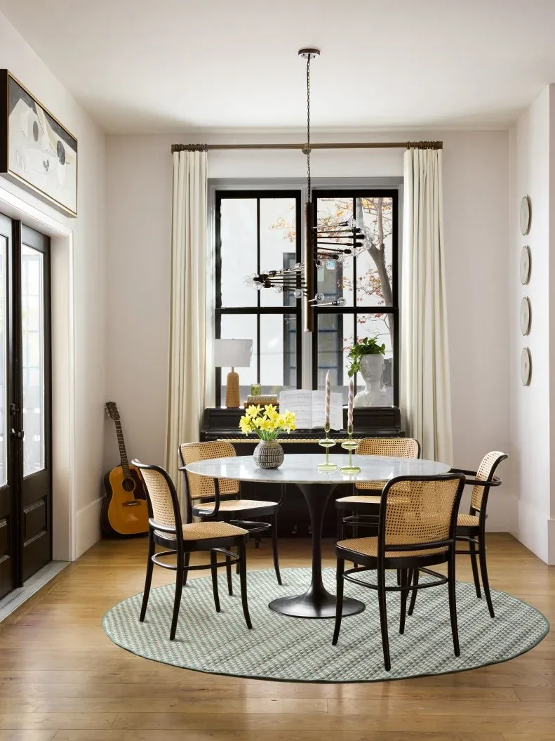 Pale full-length curtain panels on a dark rod around a black-framed dining-room window, with a round table, woven-back chairs, and a green rug.