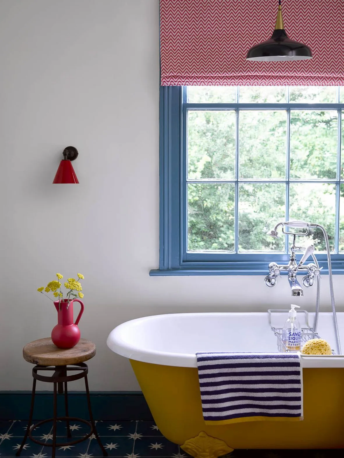 Country bathroom with a yellow freestanding bath, blue window trim, red wall light and patterned blind above a dark patterned floor.