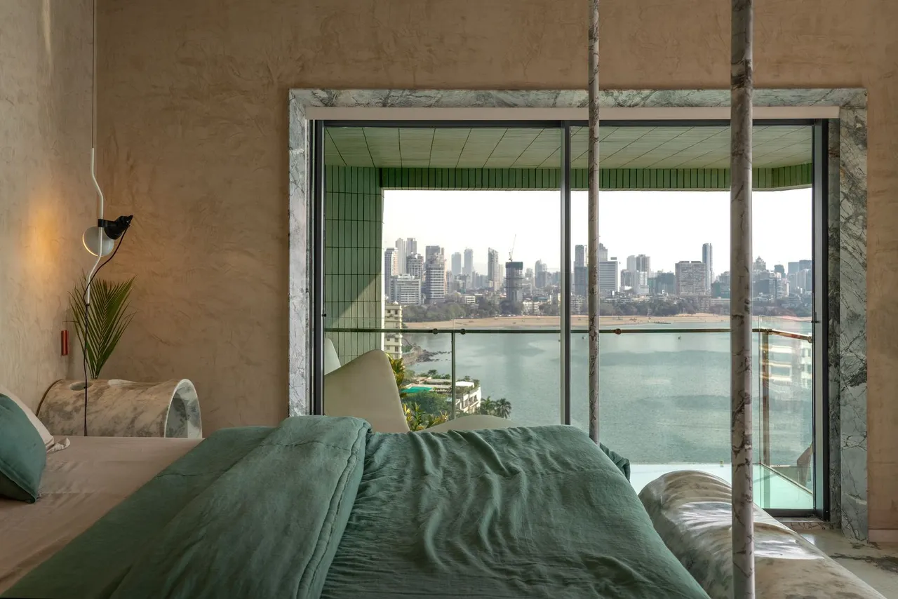 A bed with green bedding faces a broad window beneath warm neutral walls.