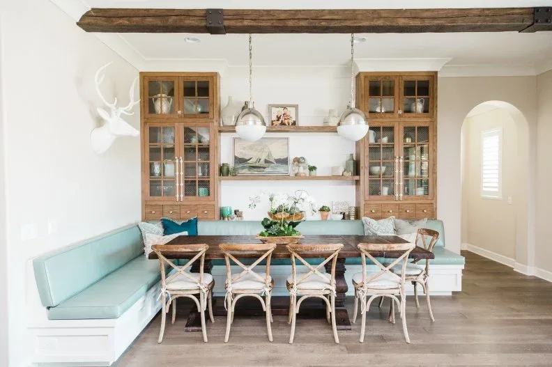 Wooden glazed cabinet towers linked by open shelves behind a pale aqua wraparound bench, with a wooden table, pale cross-back chairs, white walls, and exposed beam.