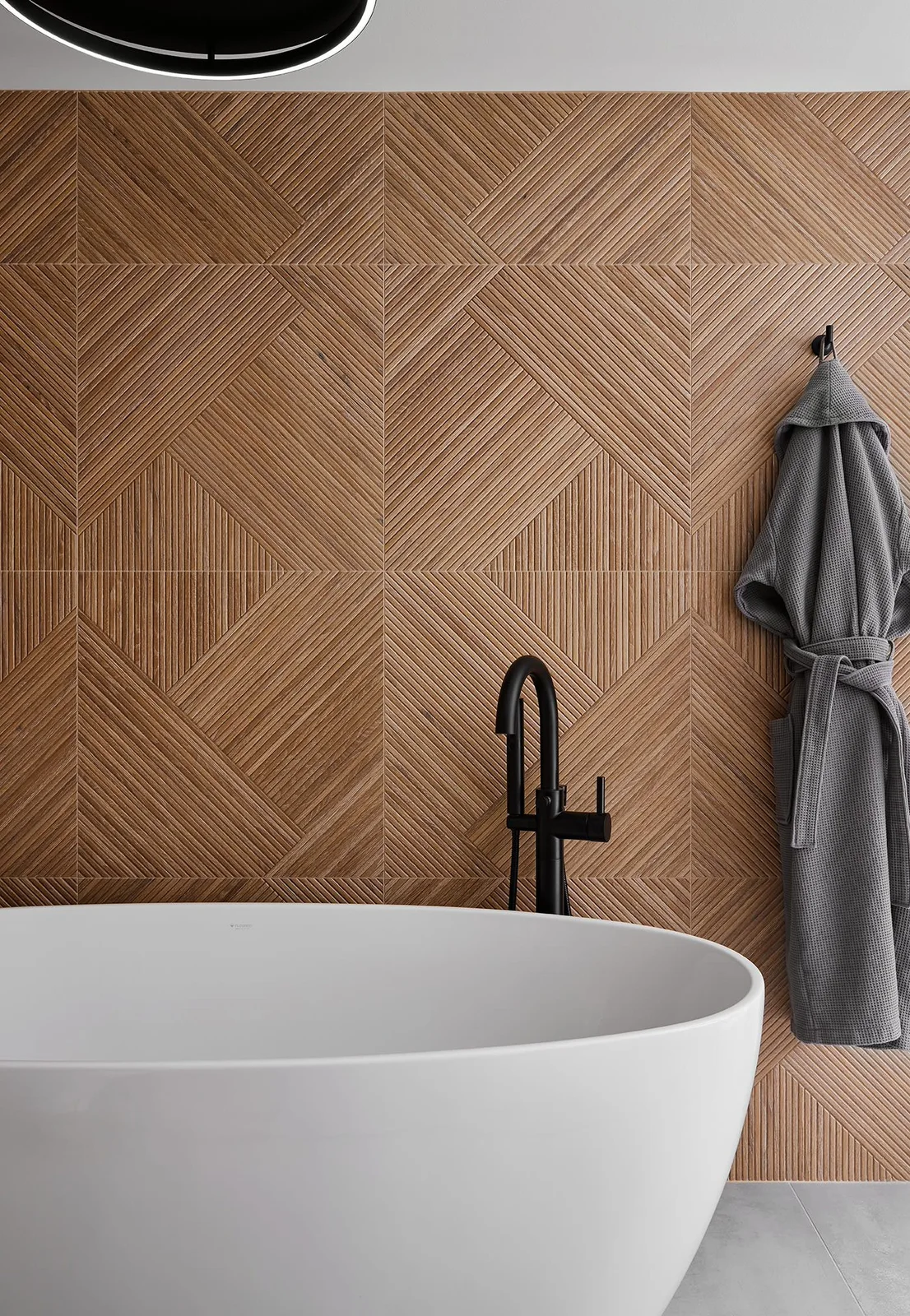 A white freestanding bath sits beside a black faucet against warm brown ribbed tiles forming geometric blocks, with a gray robe on a hook