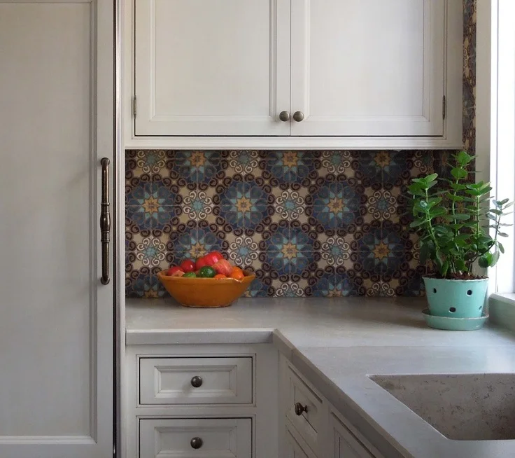 Pale cabinets surround a sink corner beneath small multicolored rosette-patterned tiles with a fruit bowl and green plant beside the window