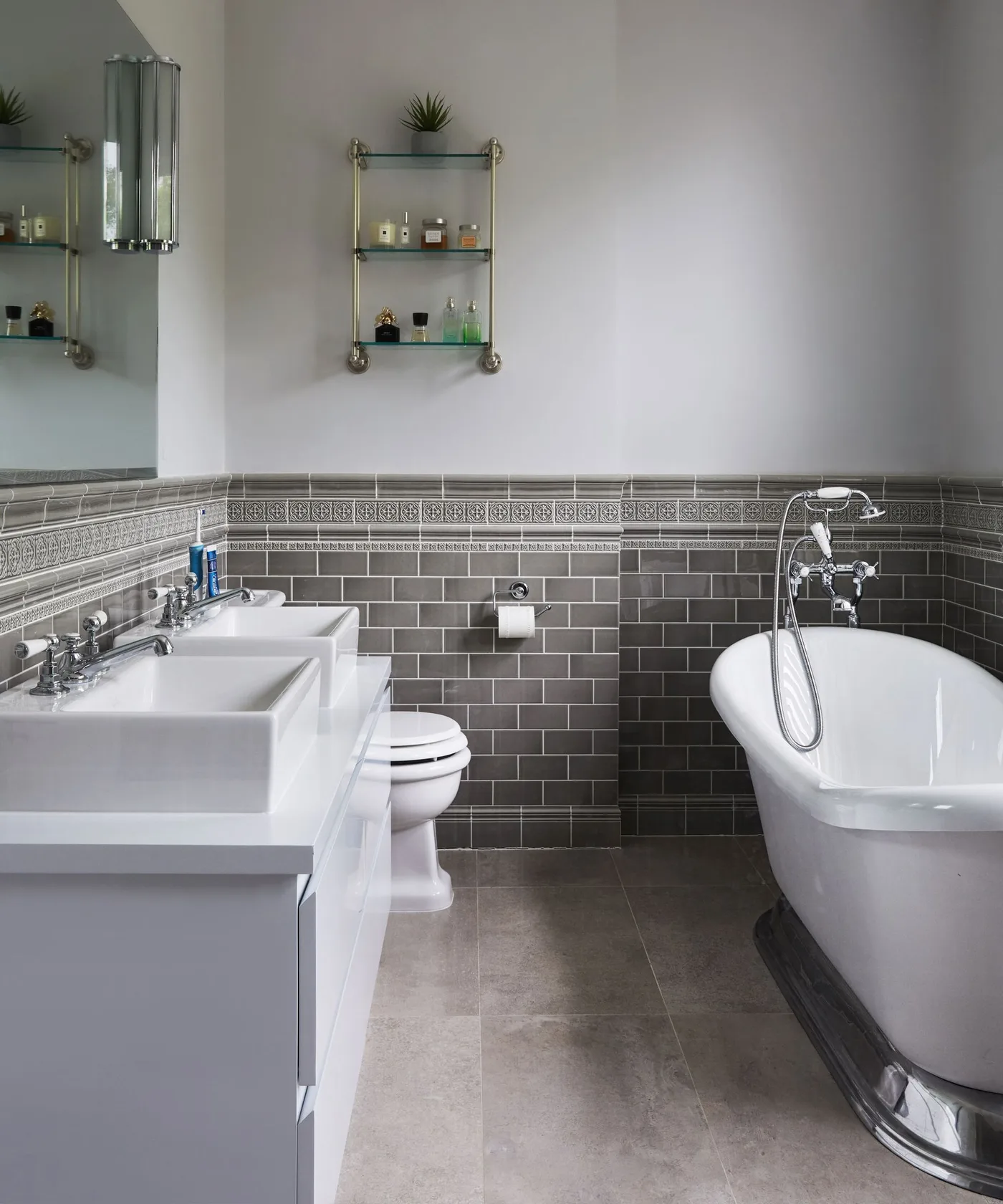 Bathroom with dark gray rectangular lower wall tiles, decorative border, pale upper walls, white square basin, slipper bath, and small glass shelves.