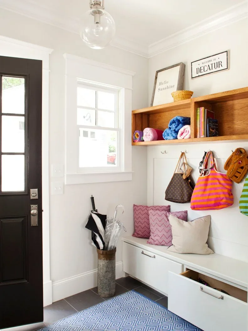 A white entry bench with drawers sits beneath a wood box shelf and coat hooks beside a dark glazed door, window and patterned blue rug