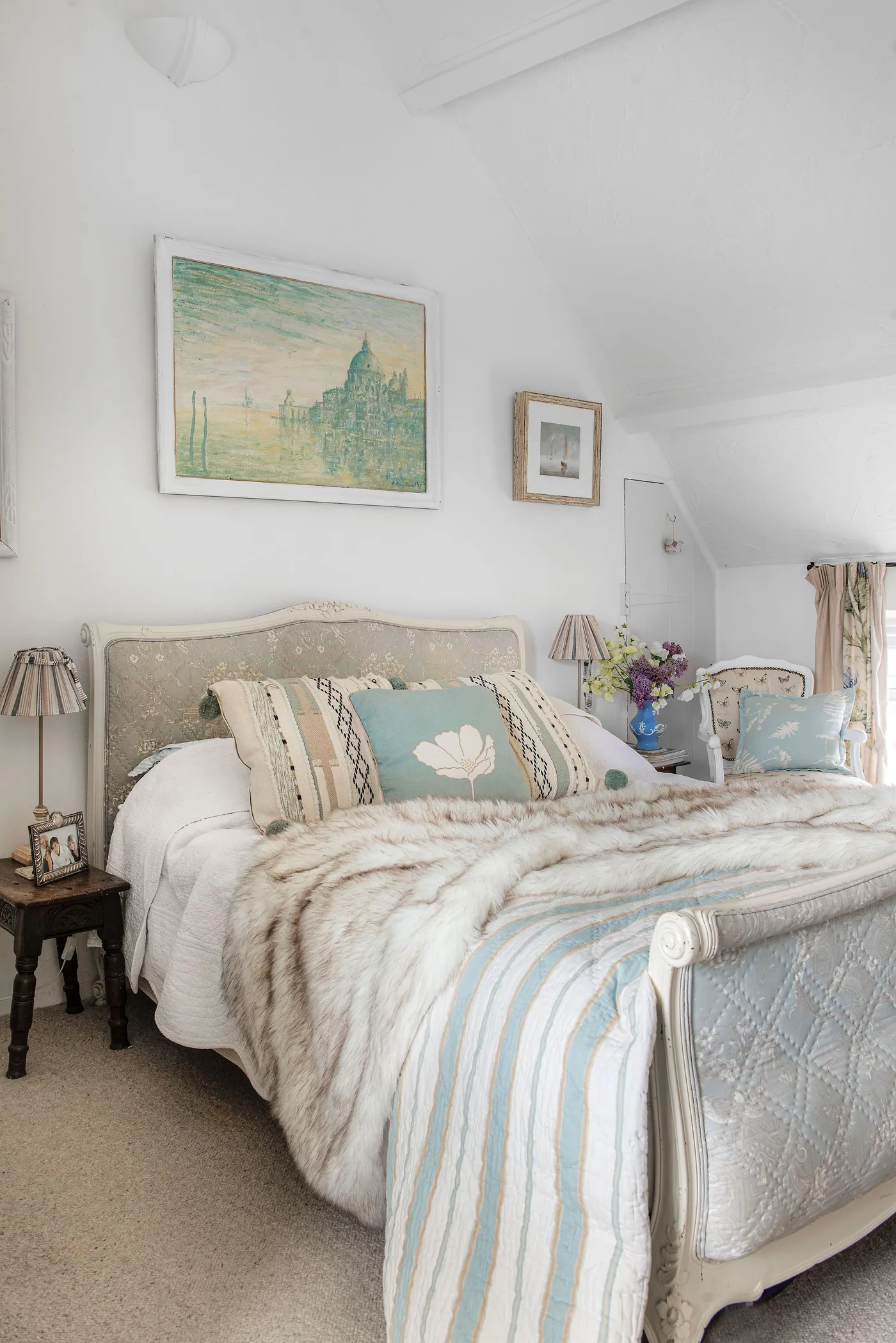 Bedroom with a pale shaped bed frame, striped cover, mixed light cushions, pleated bedside lamps and framed pictures.