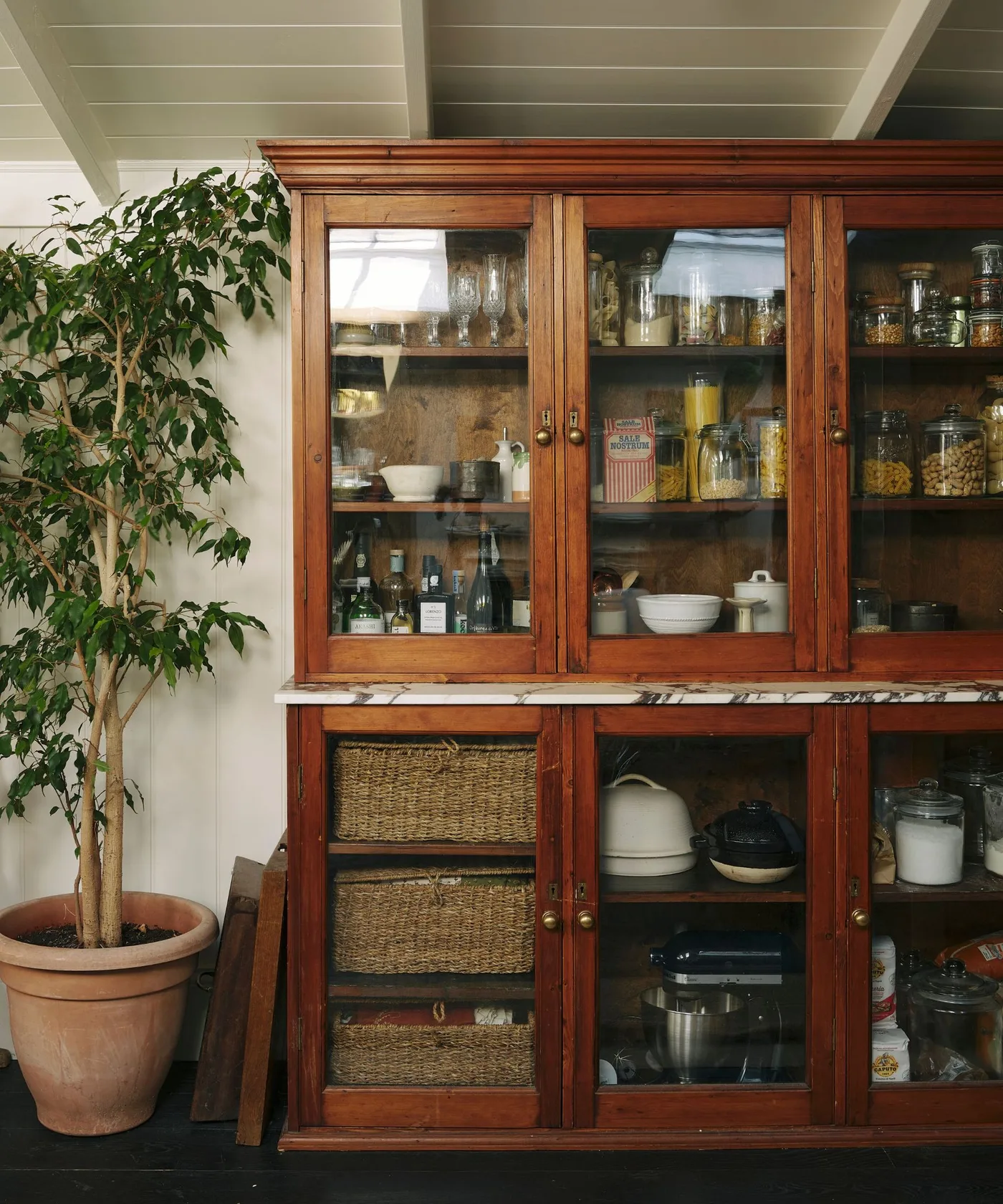 Warm wood glazed cupboard filled with jars, dishes and baskets beside pale walls and large plant