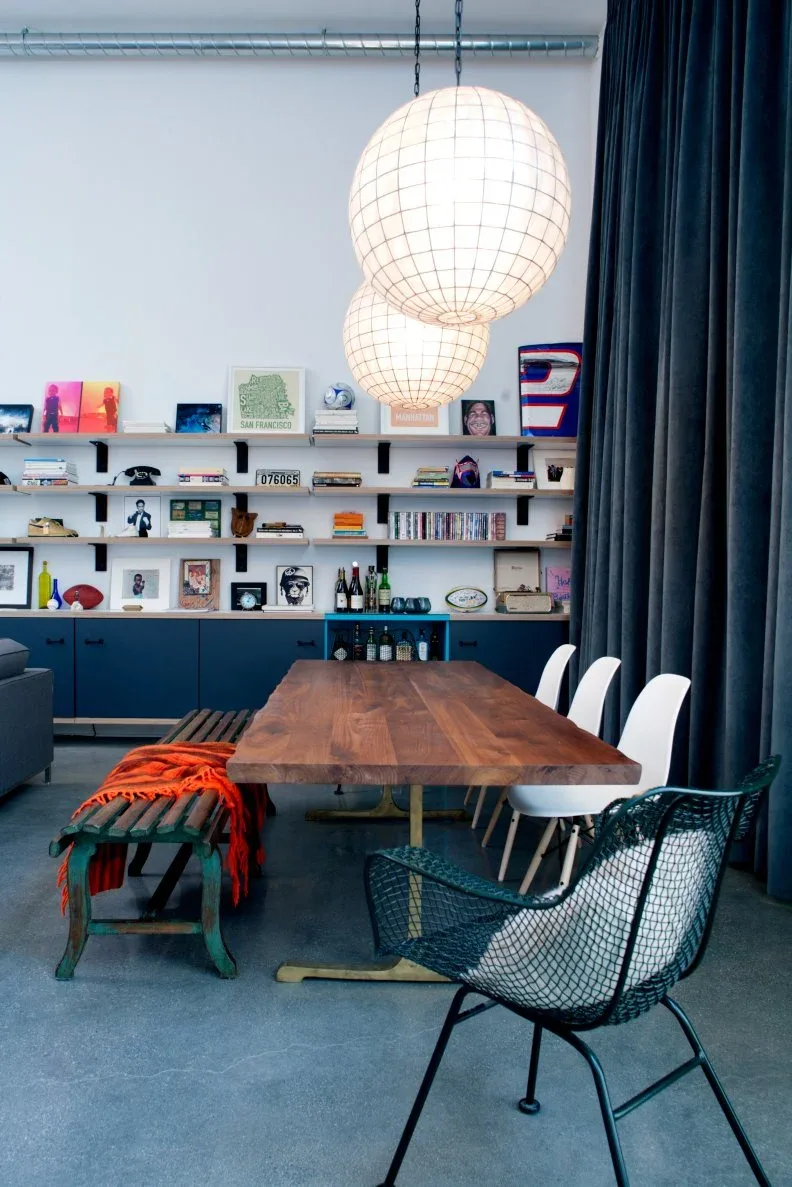 White curved dining chairs along a wooden table, with a green bench, dark open-weave end chair, blue lower cabinets, and pale globe lights.