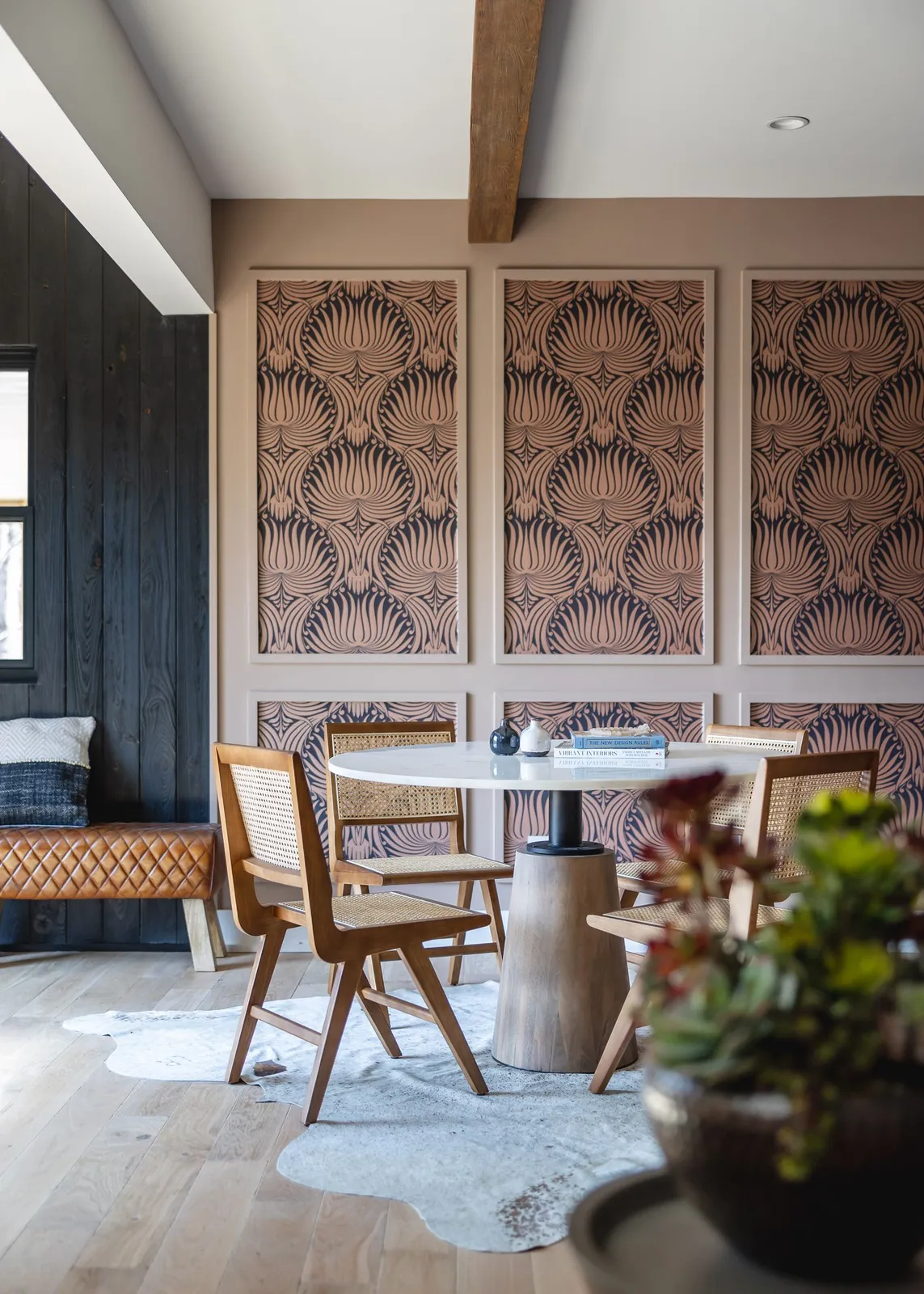 Pink-and-dark-blue patterned wallpaper fills framed wall panels beside a quilted tan bench and a pale round table with woven-back chairs