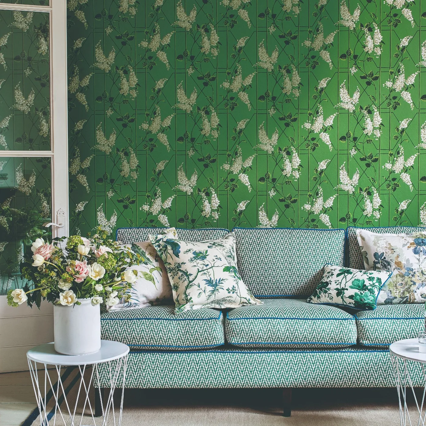 Green botanical wallpaper sits behind a green-and-white patterned sofa with floral cushions beside a glazed door