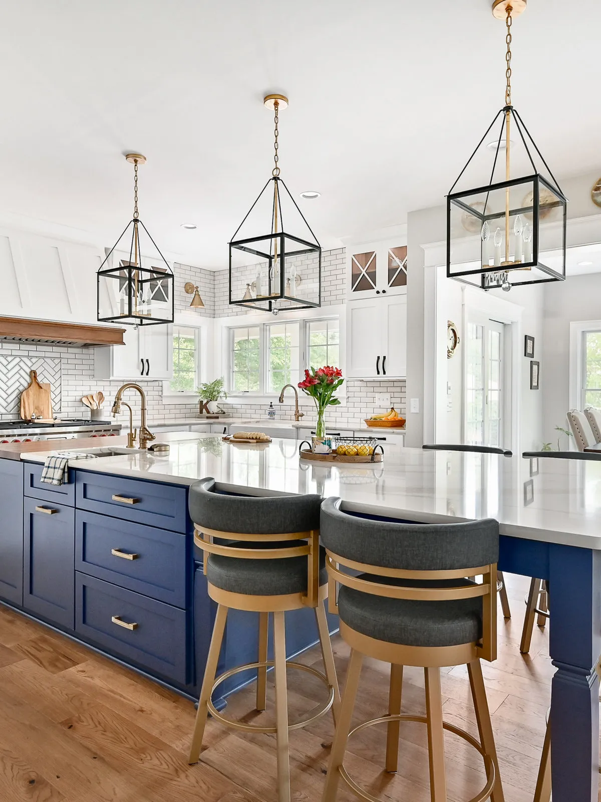 A navy island stands below open dark lantern pendants with pale counters and dark upholstered stools beside white kitchen cabinets and warm wood details