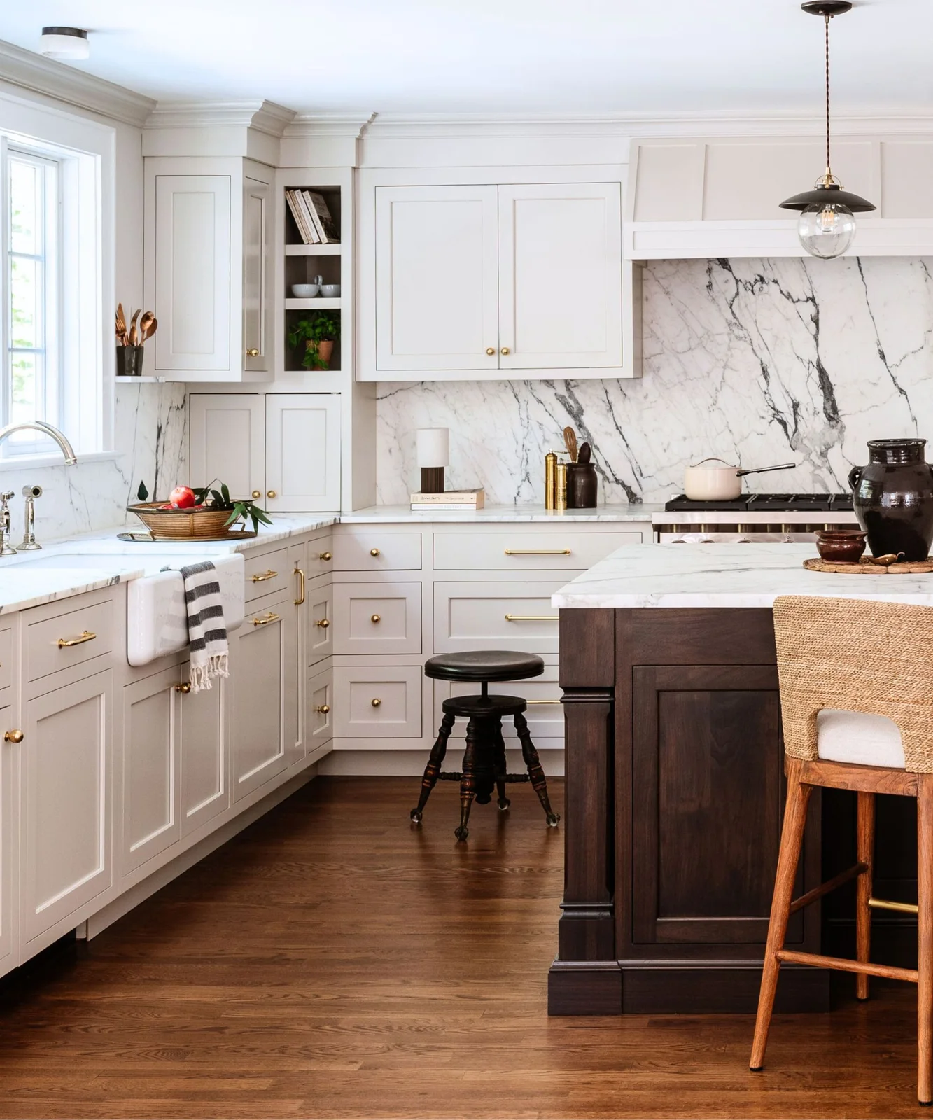 A dark wood island and woven-backed stool sit beside white framed cupboards and pale heavily veined backsplash beneath a simple globe pendant