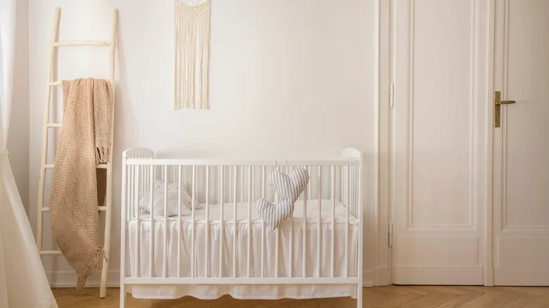 White crib with staged pillow and loose bedding against warm neutral walls beside wooden ladder and fringed wall hanging.