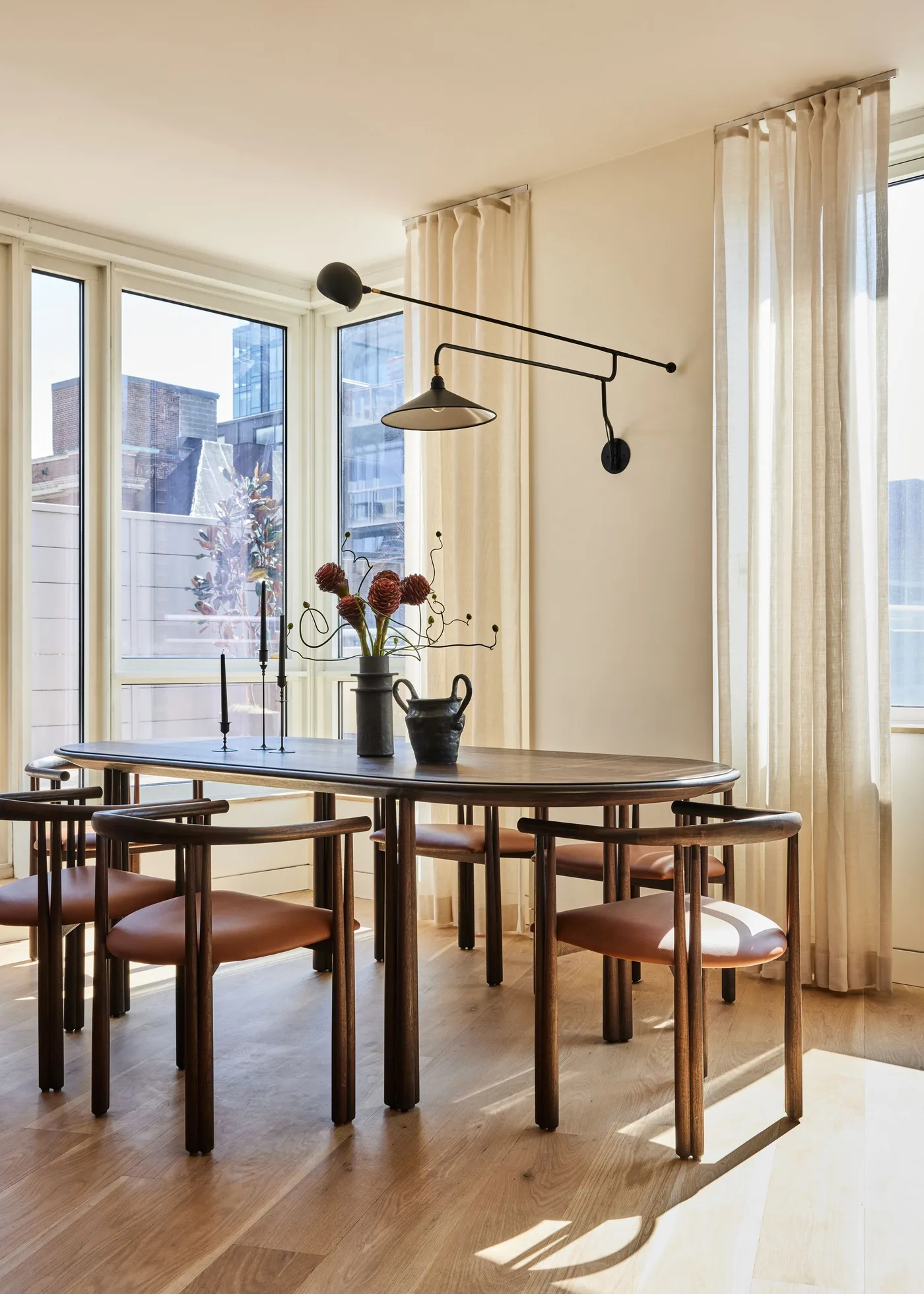 Long black wall-mounted light extending above a dark oval table beside pale curtains and tall windows