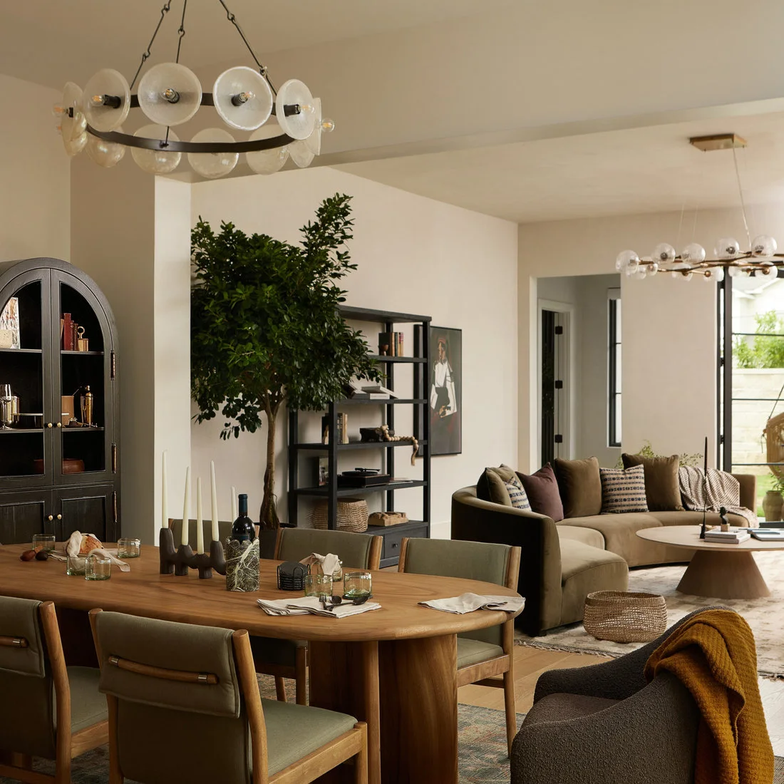 An oval wood dining table with green upholstered wood-frame chairs sits beneath a circular disc light beside a black arched cabinet, plants and a curved sofa