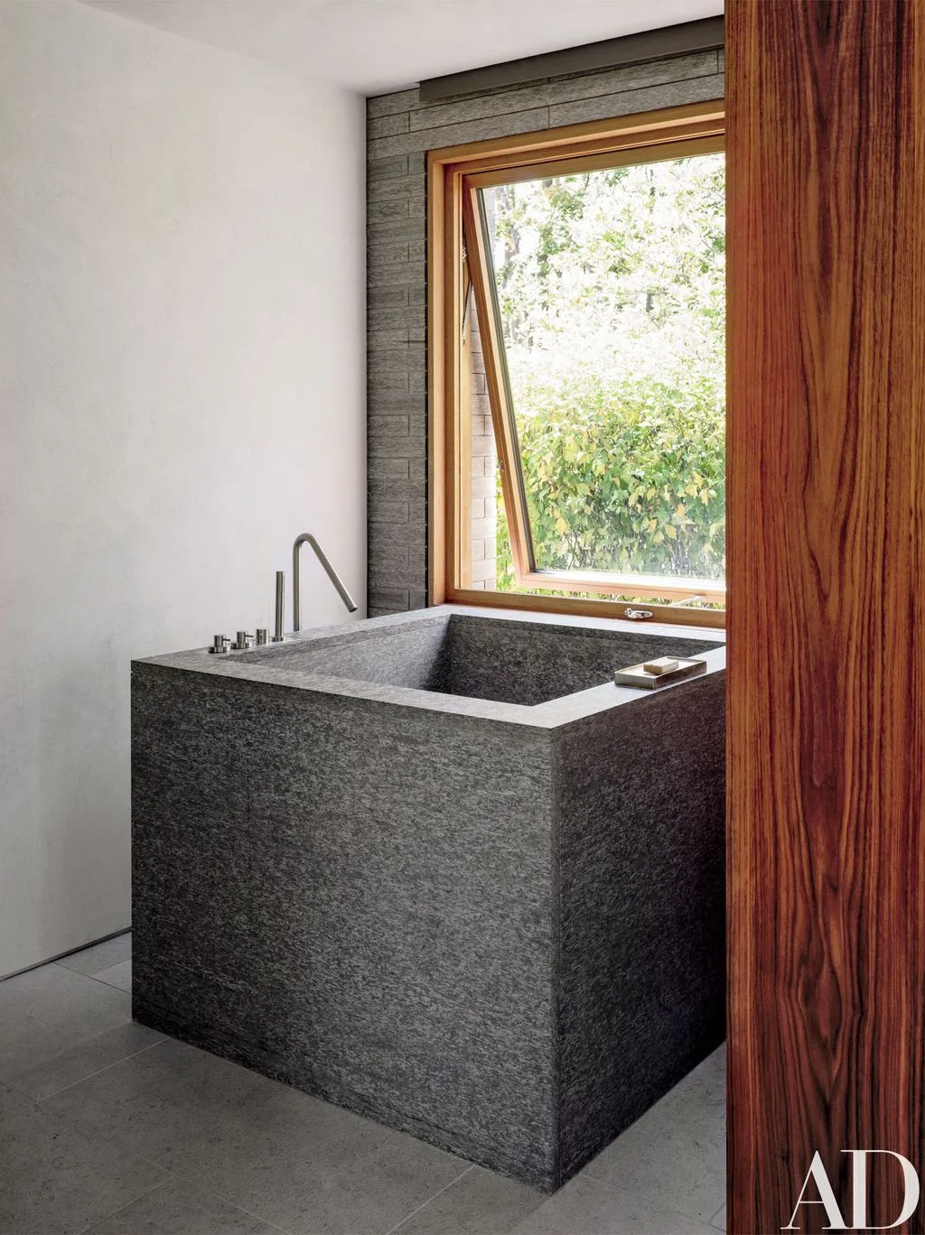 Dark speckled rectangular soaking tub beside a tall wood-framed window and wood screen, with pale walls, gray floor, and simple bath fittings.