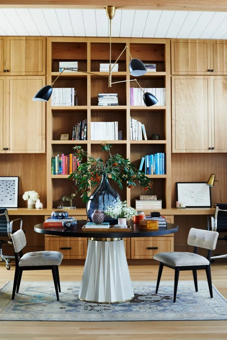 Home office with broad wooden storage wall, open bookshelves and closed cupboards, round table, pale chairs, dark pendant shades and patterned blue-gray rug.