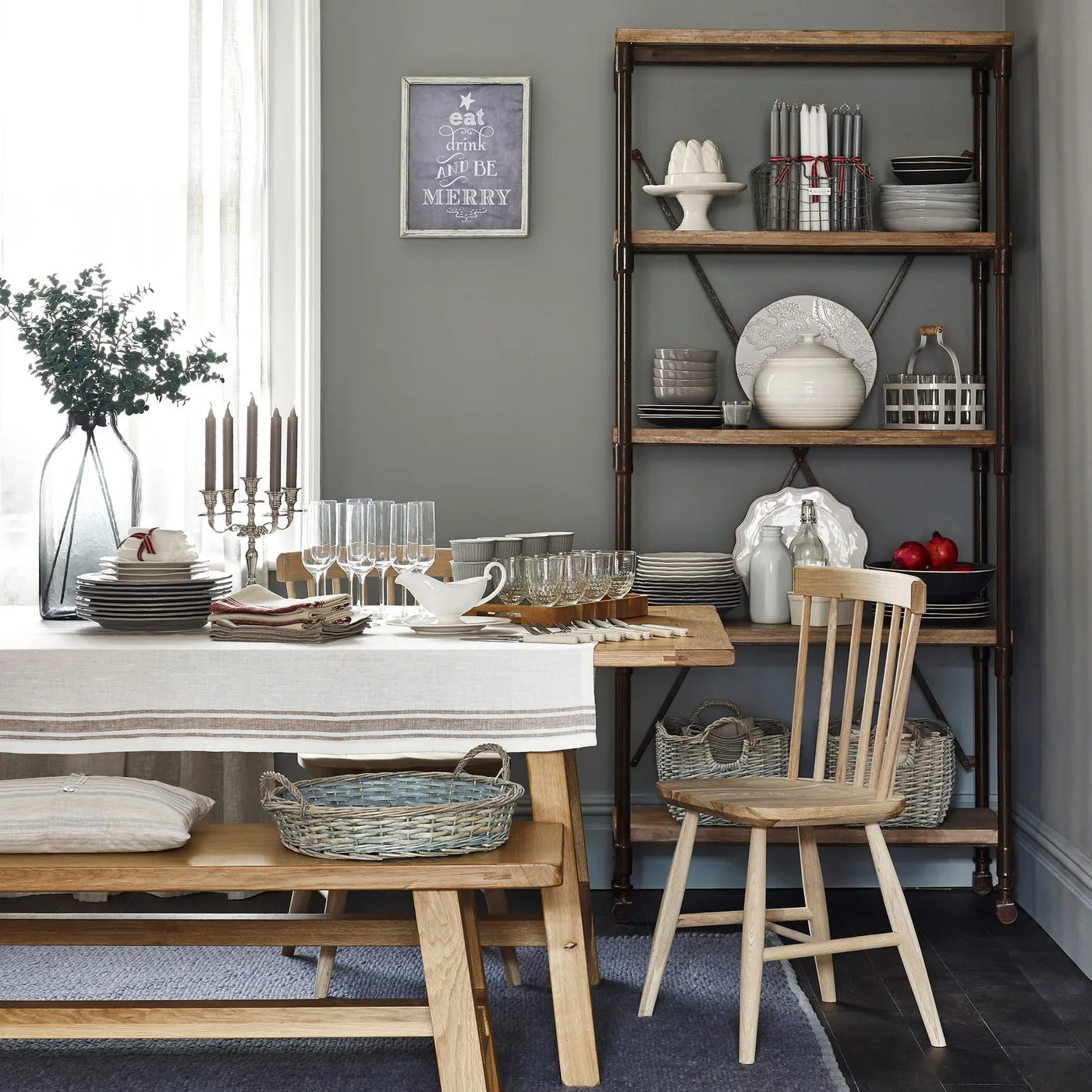 Cottage dining corner with a wooden table and bench, green-gray wall, tall open wood-and-dark-frame shelving and pale dishes.