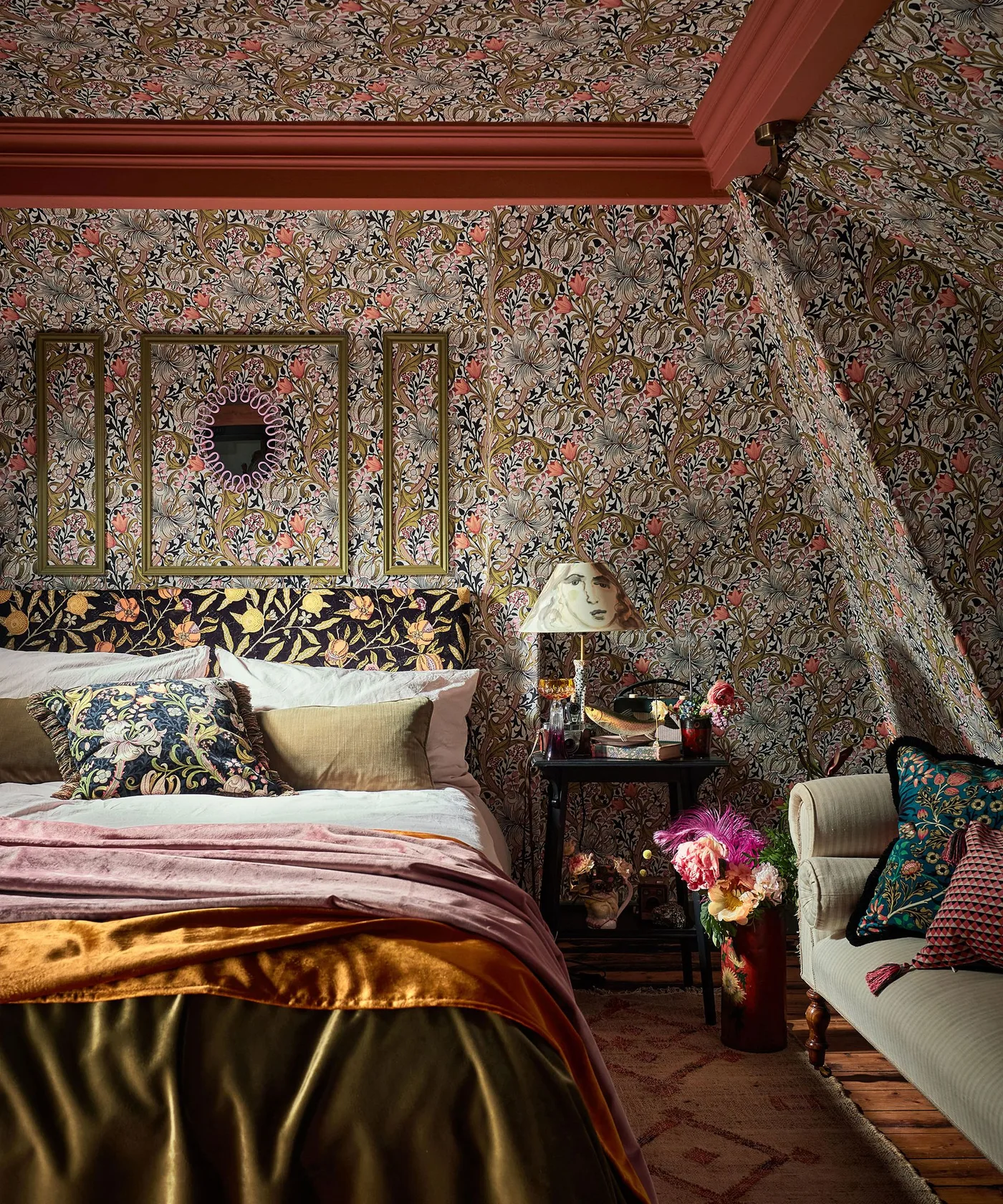 Dense olive-toned floral wallpaper on bedroom walls and ceiling with red trim and layered patterned bedding.