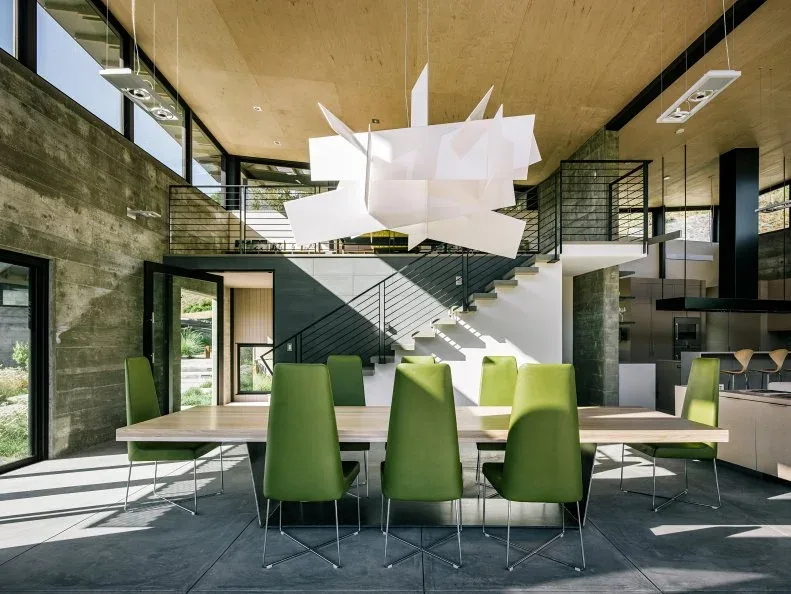 Large white angular suspended light above a long pale dining table and green chairs, with a wooden ceiling, concrete-colored walls, and open stairs.