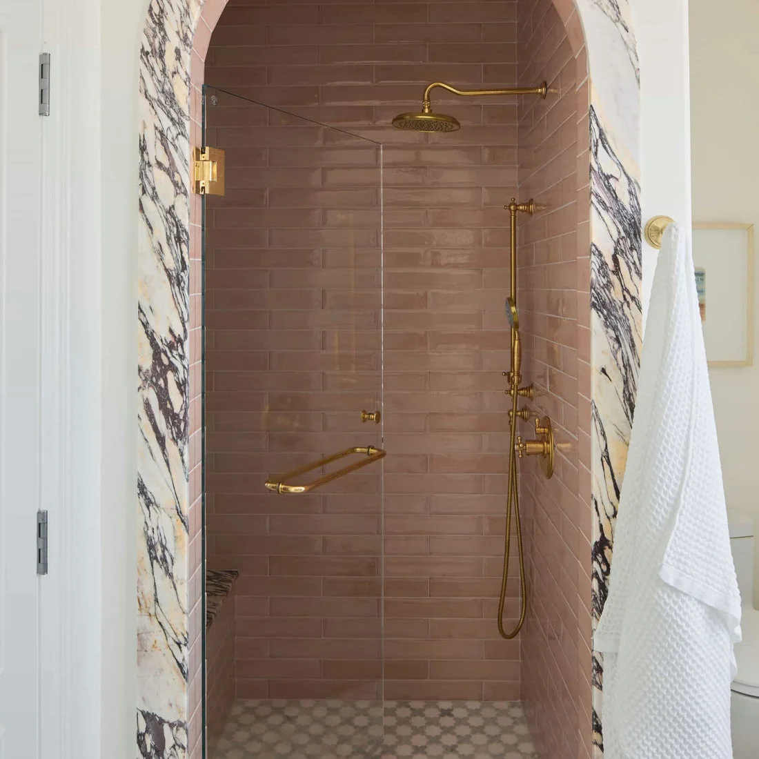 Dusty pink rectangular tiles line a shower inside a pale veined arch beside gold-colored fittings