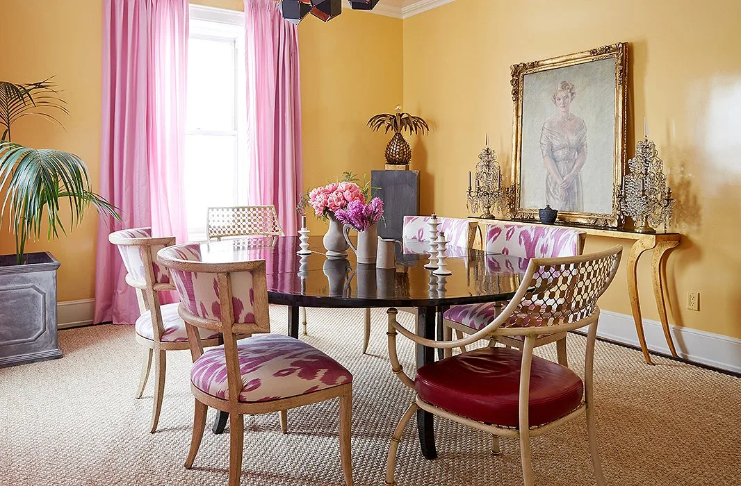 Round dark dining table with pale open-back chairs, pink curtains and yellow walls.