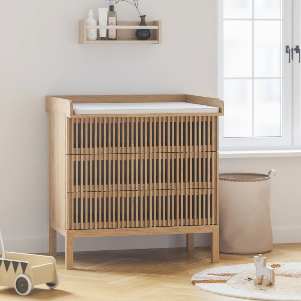 A wooden slatted changing chest with a pale pad stands beside a window and a small wall shelf