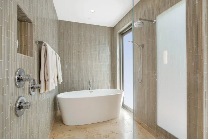 A white oval bath stands beside a clear shower enclosure within narrow warm-toned wall tiles, with a recessed shelf, towels and a glazed outer wall