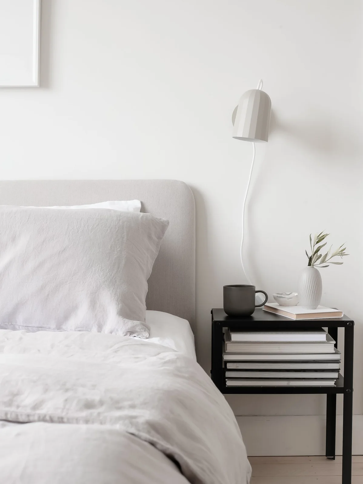 A white bed and pale curved headboard sit beside a black open bedside table holding books below a white wall lamp