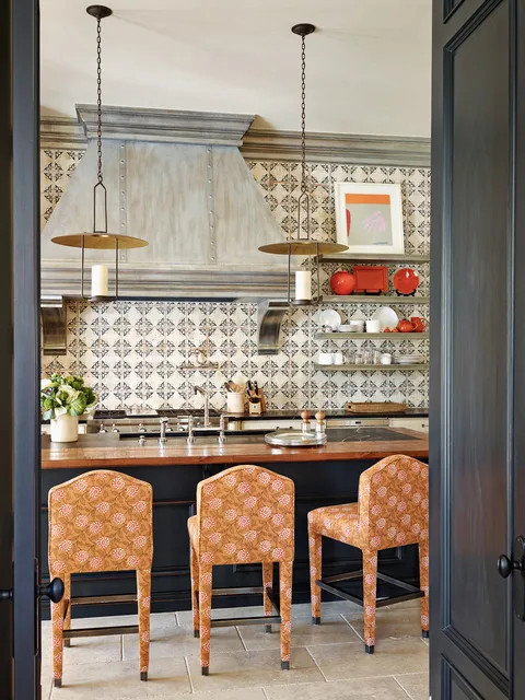 Dark cabinetry frames a small kitchen with a patterned backsplash and warm upholstered stools.