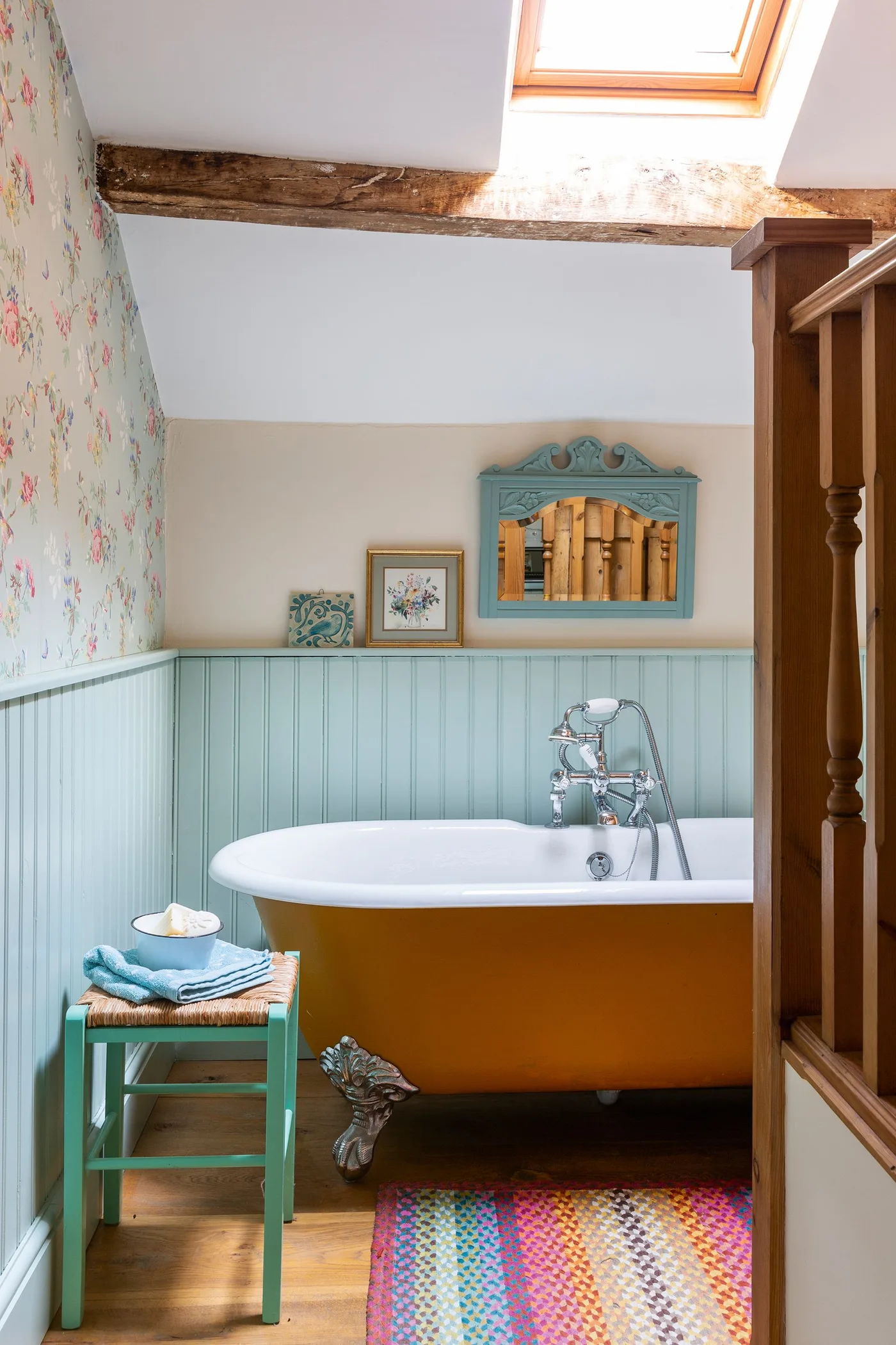 Bathroom with an orange freestanding bath, pale blue vertical paneling, floral wallpaper, blue framed mirror and a skylight above a beam.