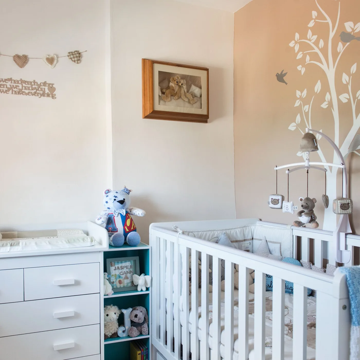 White crib beside pale tree mural, wooden-framed picture, small garland and blue-green open storage.