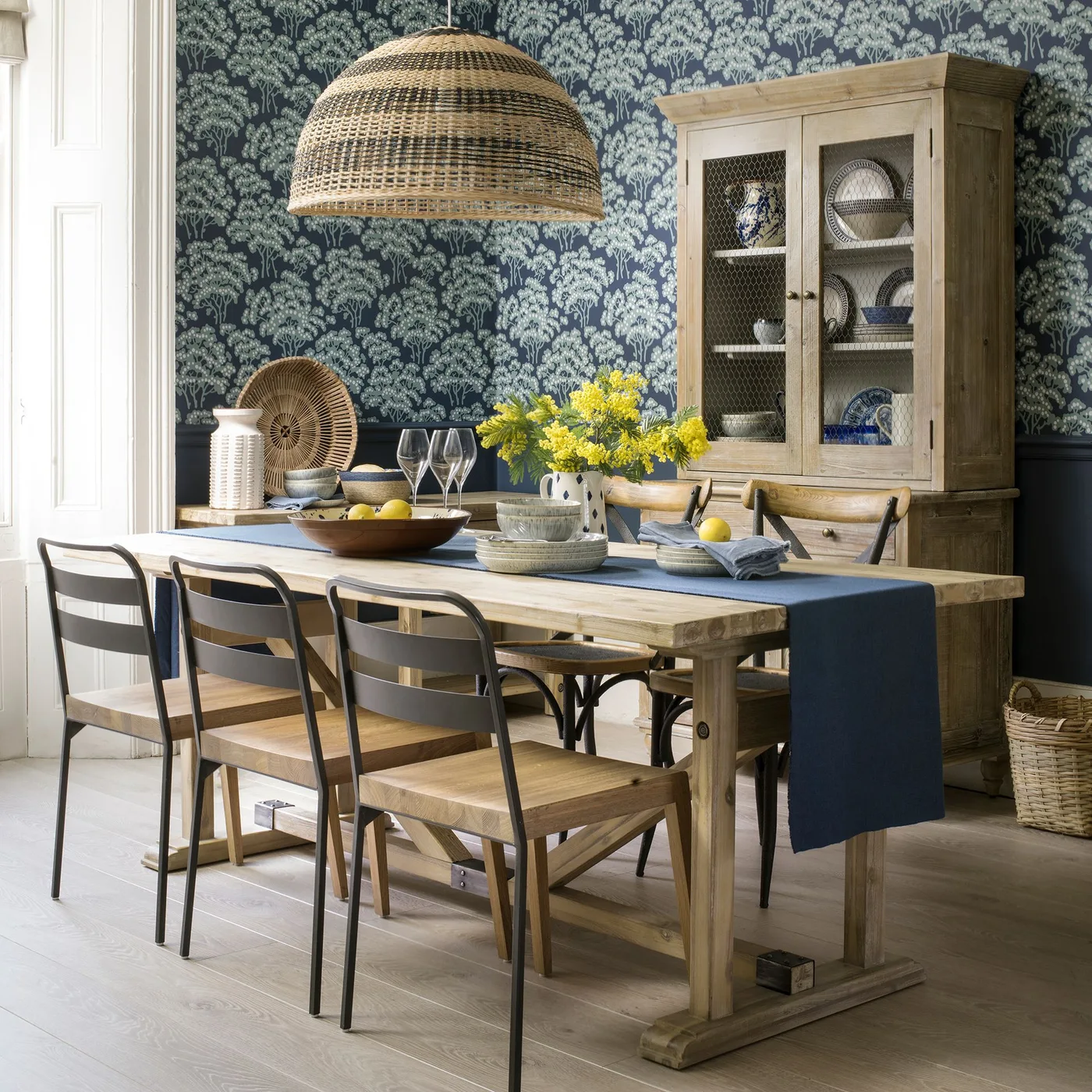 Cottage dining room with dark blue leafy wallpaper, wooden table, mixed wooden and dark-framed chairs, tall dish cupboard and woven pendant shade.