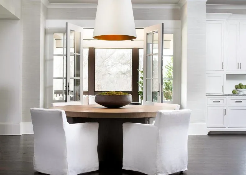 Broad dark bowl with a low green arrangement on a round wood dining table beneath a white pendant