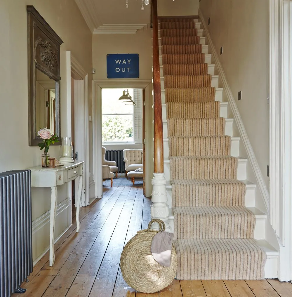 Pale hallway with a striped stair runner, slim white console, dark radiator and warm wooden floorboards.
