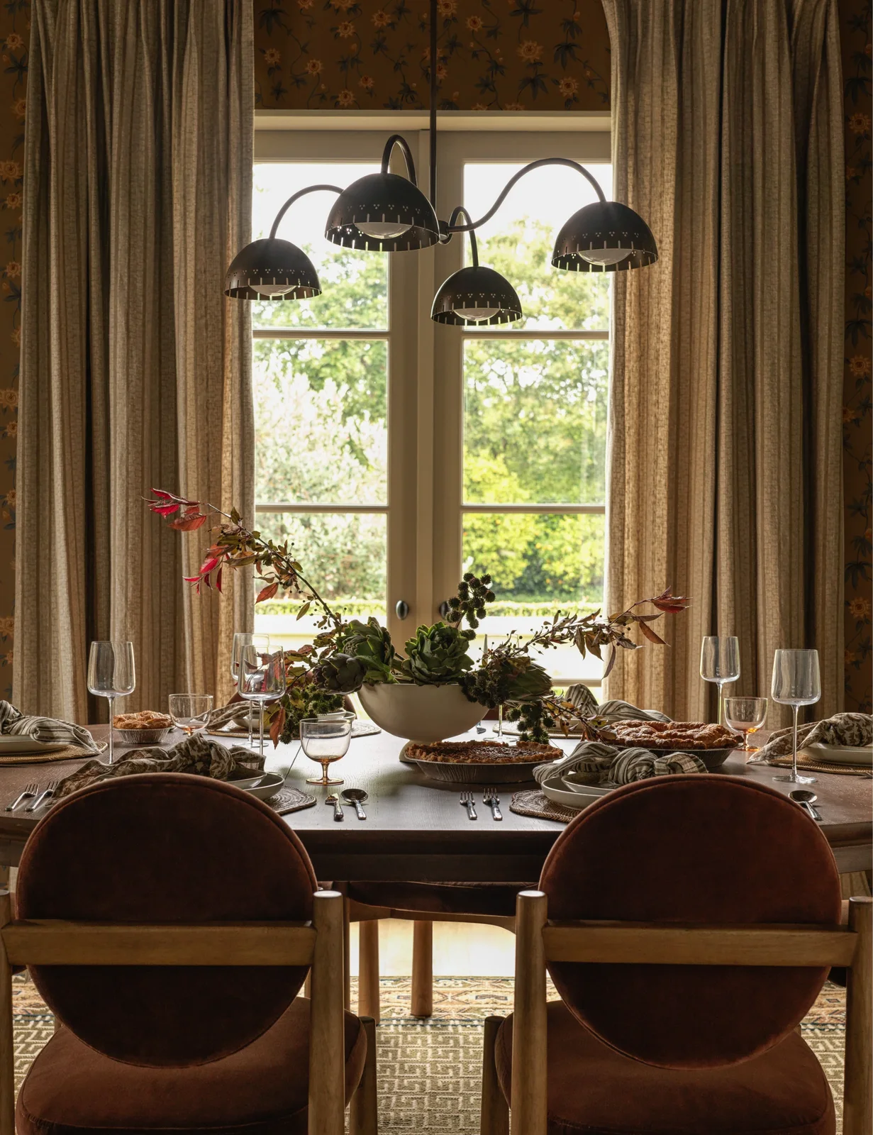 A pale bowl holding green artichokes and leafy branches sits on a dark dining table beside clear glasses and neutral napkins