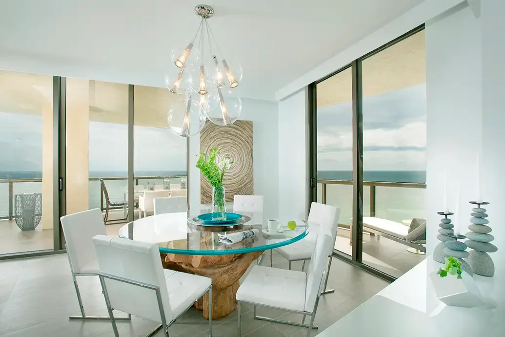 White dining chairs stand around a round clear glass table with a sculptural wood-toned base beside tall dark-framed windows and turquoise table accents