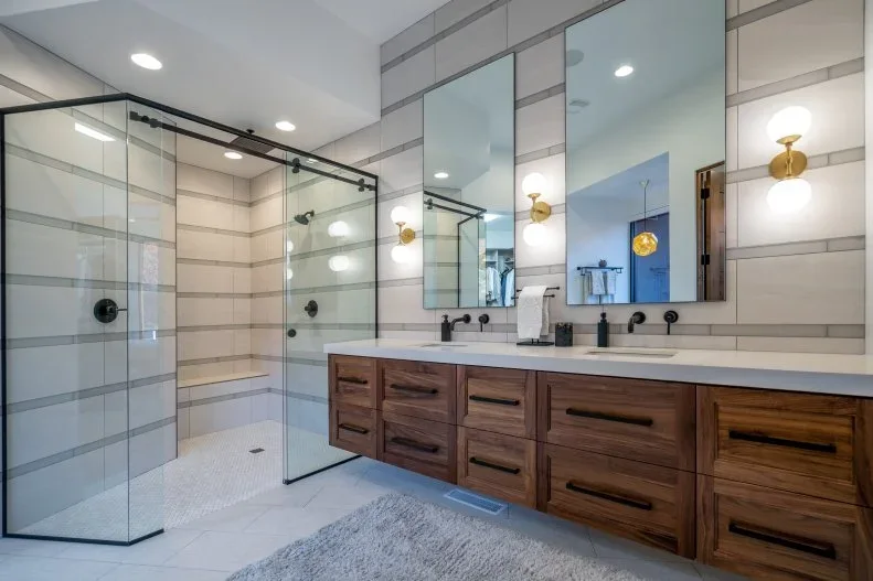 A long wood drawer vanity with paired basins stands beneath separate mirrors beside a clear dark-framed shower enclosure within pale horizontal tile bands