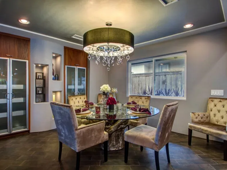 Wooden fitted cabinets with glazed doors and small open niches beside a gray dining area, pale upholstered chairs, a glass table, and a shaded chandelier.